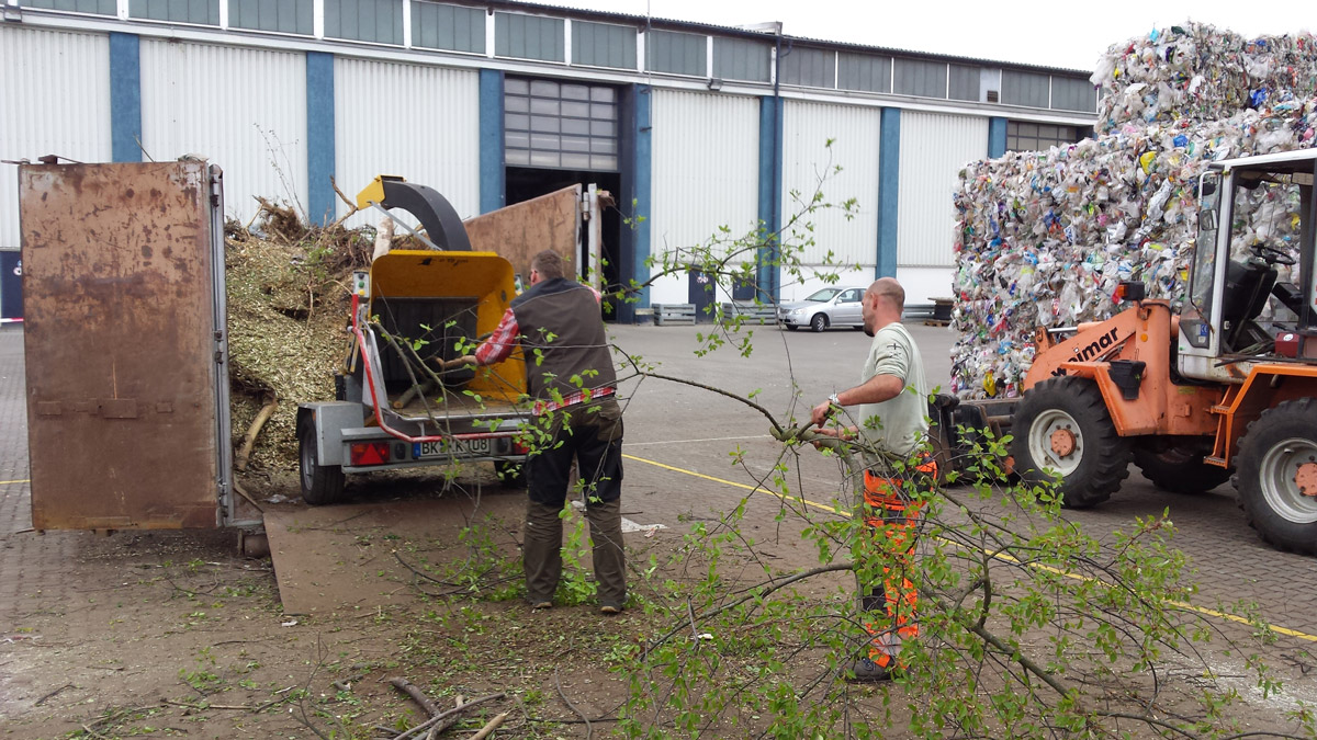 Zwei Männer in Arbeitskleidung, die auf einem Firmengelände Grünschnitt in einen mobilen Container häckseln.